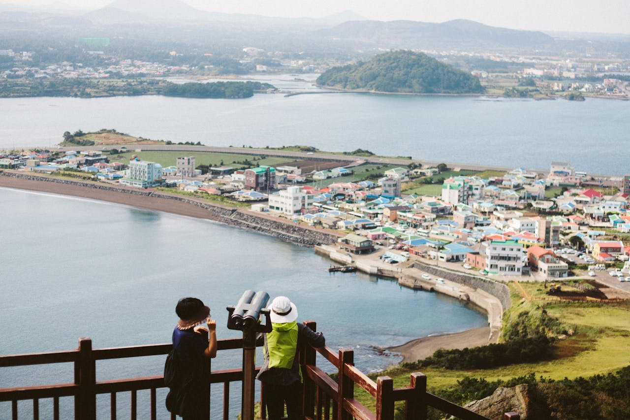 Tourists enjoying a panoramic view from Seongsan Ilchulbong overlooking Jeju Island and coastal village.